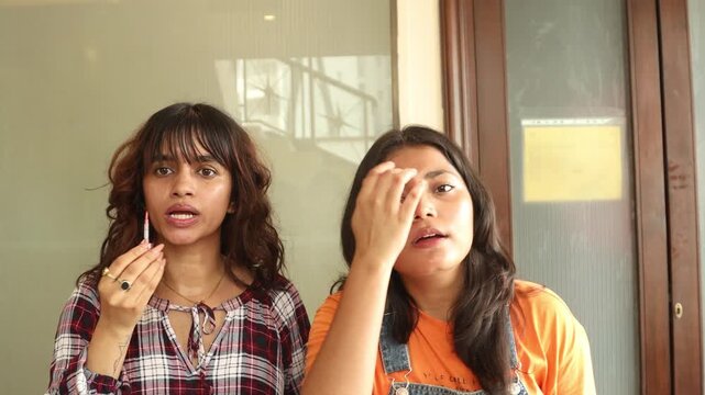 Two young indian Gen Z female friends applying lipstick together, sharing a casual beauty and makeup routine, self-care, cosmetics, personal grooming, youth lifestyle, and modern beauty culture.