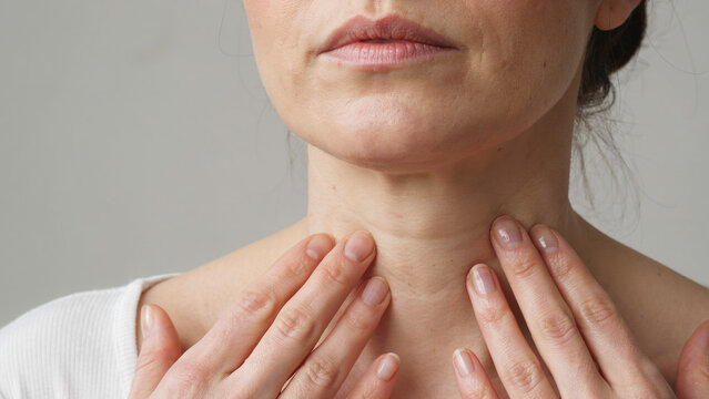 Woman's hands gently touching her neck and throat area in close-up against a light background.