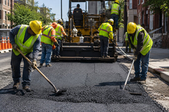 Road construction crew wearing safety vests and hard hats laying and leveling fresh asphalt paving on a city street for infrastructure projects