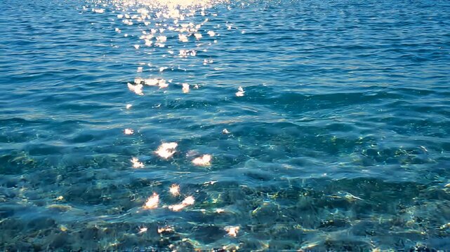 Blue sea surface with sun reflections on gentle ripples revealing pebbles beneath the shallow calm ocean.