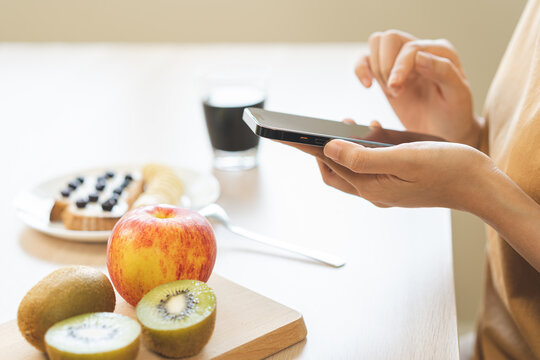 Asian young woman using smartphone or mobile for take a photo while having healthy breakfast with fruit toast, kiwi, apple and black coffee on table, calorie counting or morning lifestyle concept.