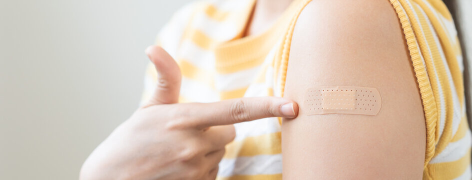 Close up of a young woman pointing at an adhesive bandage on her arm after receiving a vaccine injection, representing healthcare, virus immunity protection, and prevention.
