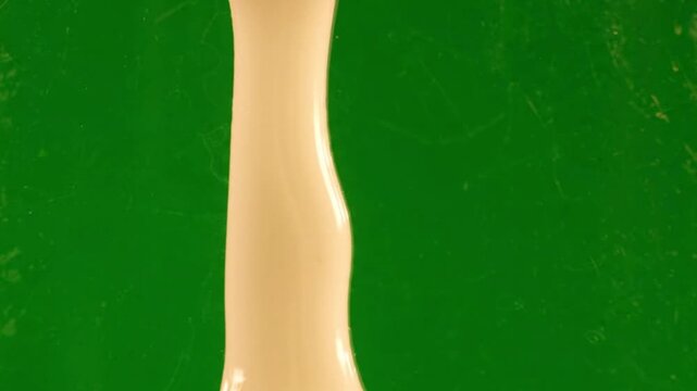 Thick beige liquid dripping slowly, close-up shot of cream substance flowing downwards against a solid green background, concept of wax, paint or cosmetic cream movement