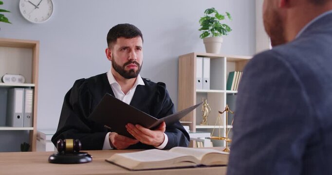 Confident man judge wearing black judicial robe knocking gavel having court verdict sitting at the desk on workplace in courtroom with male client. Legal law and justice concept. 4k video.