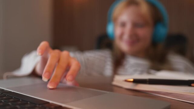 Close-up on a young student's hand using a laptop touchpad for scrolling and clicking, with the girl smiling and wearing headphones for an e-learning session in a softly lit room