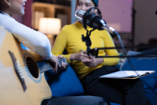 Woman musicians recording a podcast interview in studio. Two Asian women creating digital content, one playing a guitar while being interviewed by a host in a podcast studio.