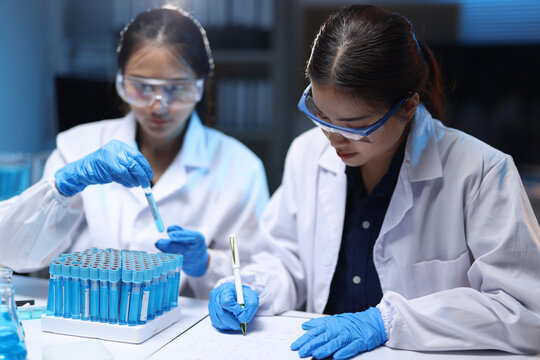 Female scientists conducting research and experiments in laboratory. Two female scientists wearing lab coats and safety glasses conducting scientific research, holding a test tube and writing notes.