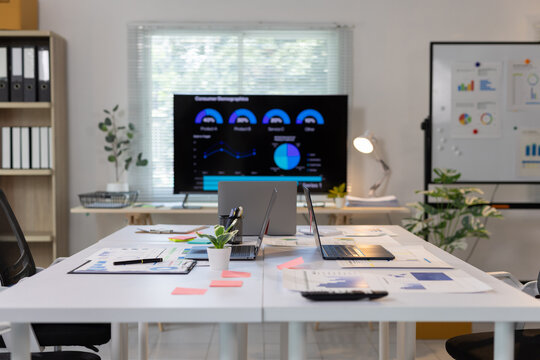 Meeting room with a large screen displaying charts and graphs, laptops, and various documents for business data analysis.