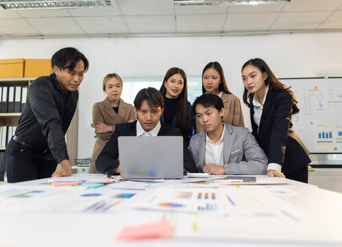 Asian business team collaborating on financial data analysis. Diverse Asian business team working together, analyzing reports and charts on a laptop and physical documents during an office meeting.