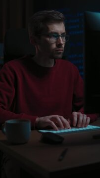 Male coder typing programming script on a keyboard late at night, code reflecting in his glasses. Developer works from a dark home office, concentrated on debugging . Vertical video