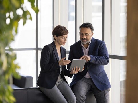 Professional business partners reviewing information on a tablet in a sunlit office, male and female managers working together for successful startup growth and communication.