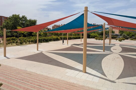 Parasoles triangulares de exterior, de color rojo y azul, sostenidos por postes de metal en una plaza p&uacute;blica