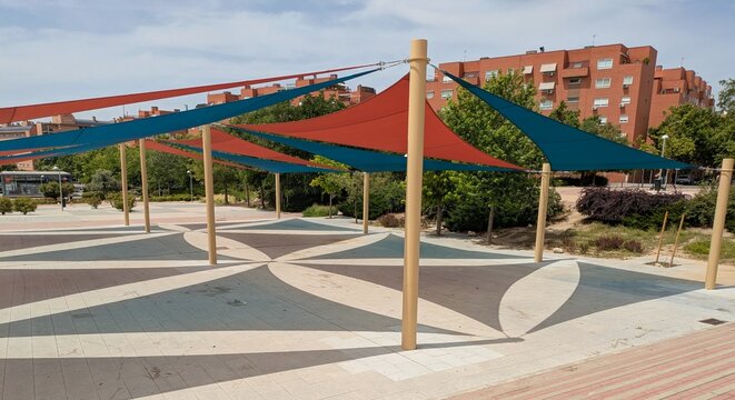 Parasoles triangulares de exterior, de color rojo y azul, sostenidos por postes de metal en una plaza p&uacute;blica