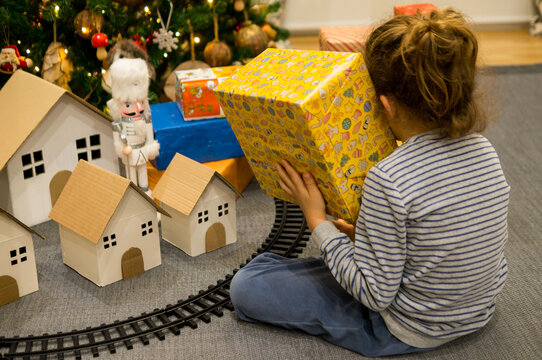 A child sitting beside toy train tracks shaking a yellow wrapped Christmas box near her ear to hear what is inside.