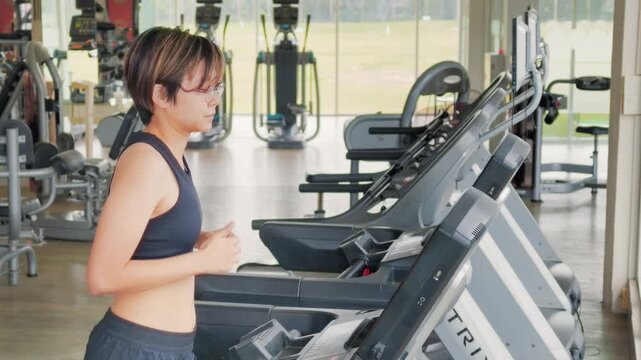Active Asian woman in her forties wearing glasses and a black sports bra runs on a treadmill inside a modern brightly lit gym. She is focused on her cardio workout to maintain a healthy lifestyle.