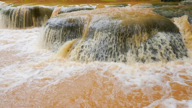 Sediment rich floodwater pours rapidly over a tropical rainforest waterfall, reflecting severe rainfall, global warming, climate instability, soil erosion, watershed stress, natural disaster danger.
