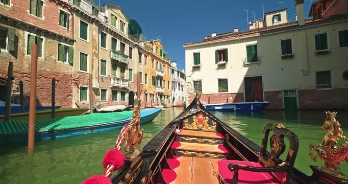 POV shot from a luxury carved gondola floating along a sunlit Venetian canal junction flanked by weathered rustic red brick houses and moored boats.