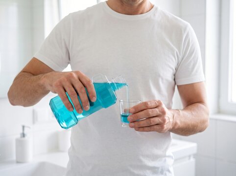 Man pouring blue mouthwash into a small plastic cup in a bright bathroom