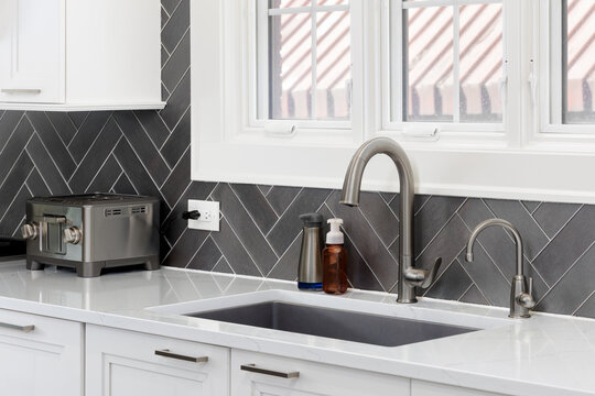 A kitchen faucet detail with a stainless steel faucet on a white marble countertop, white cabinets, and a dark grey textured herringbone tiled backsplash with white grout.