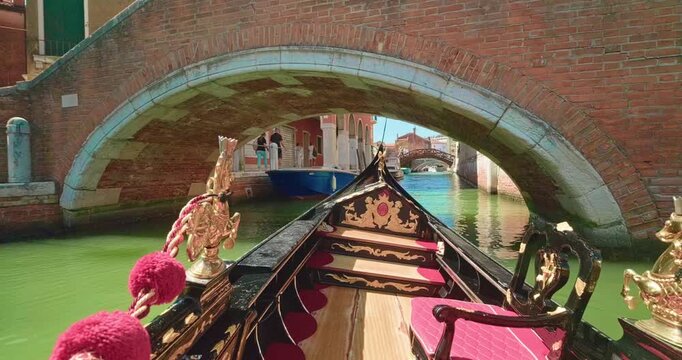 Cinematic POV shot from a traditional decorated gondola cruising along a narrow water canal and passing under an old stone bridge between historic houses in Venice.