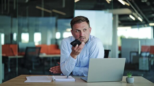 Businessman in blue shirt sends voice message on smartphone in modern office.
