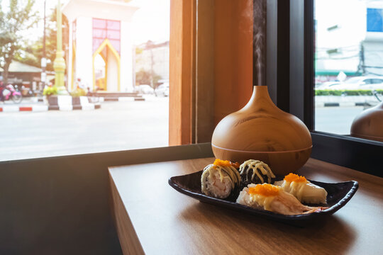 Sushi dish on window counter bar with ultrasonic humidifier. Assorted Japanese sushi, including nigiri and gunkan topped with roe next to wood grain aroma diffuser oil diffuser by window view.