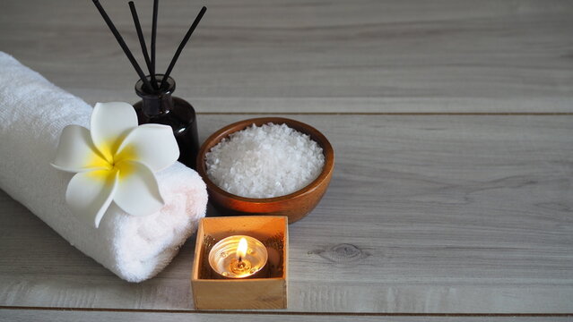 Spa wellness arrangement featuring a rolled white towel decorated with a white plumeria flower, a lit tealight candle in a wooden box, a wooden bowl filled with white bath salts, and a reed diffuser b