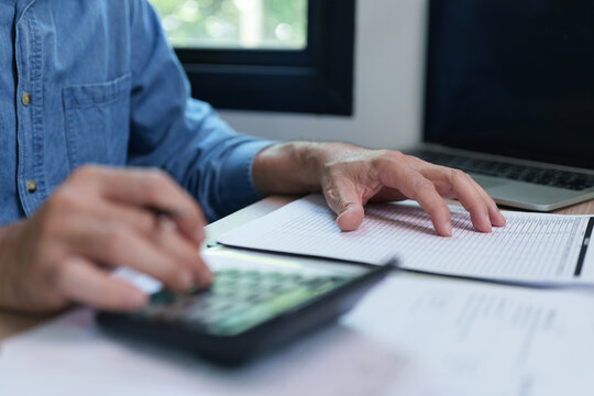 Man using a calculator while reviewing financial reports and paperwork at home, representing budgeting, accounting, tax calculation, debt management, expenses and personal finance planning.