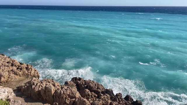 Blues waves crash against the rocks, creating a vivid and energetic coastal scene. The bright sky and deep blue sea blend beautifully of the Mediterranean Sea near Nice, Cote d'Azur, southern France.