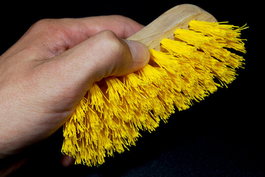 A man's hand holds a handheld surface-cleaning brush with a wooden handle and bright yellow synthetic bristles