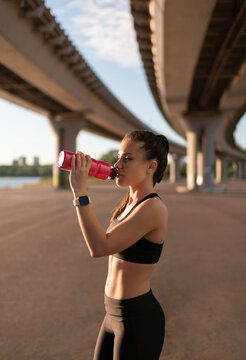 Slim sportswoman drinking water on shore under bridge