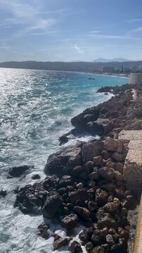 Blues waves crash against the rocks, creating a vivid and energetic coastal scene. The bright sky and deep blue sea blend beautifully of the Mediterranean Sea near Nice, Cote d'Azur, southern France.