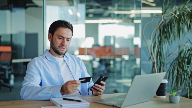 Stressed bearded businessman wearing blue shirt looking worried while holding credit card and smartphone, experiencing failed transaction error in modern office.