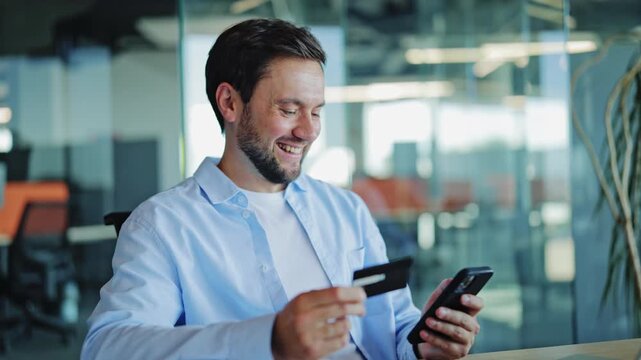 Man in blue shirt completes online payment with smartphone and credit card, smiling with satisfaction in a modern office.