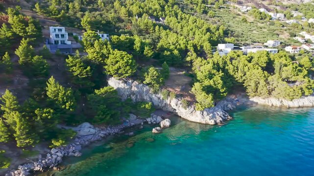 Aerial drone view of a rocky Adriatic Sea coastline with crystal clear turquoise water, pine trees, white stone cliffs, secluded coves and coastal houses.