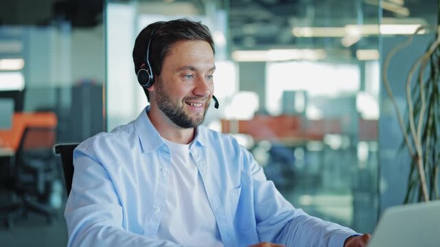 Male support specialist wearing a light blue shirt and a headset actively works on his laptop. Man provides excellent customer service in a contemporary open-plan office setting.