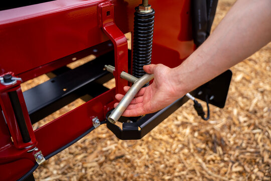 Hand holding metal T-shaped adjustment handle near heavy spring on red frame of industrial machine.