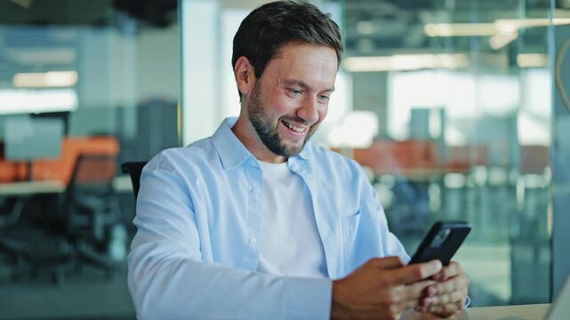 Businessman with a beard and light blue shirt smiles and celebrates good news while looking at his smartphone in a bright, modern office environment.
