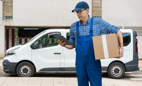 A courier holding a cardboard box and digital tablet with a delivery van in the background.