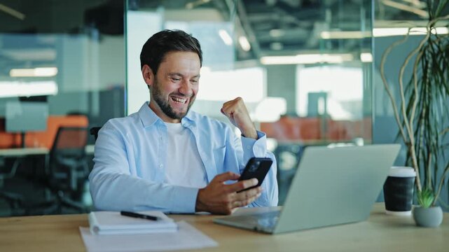 Male executive in a light blue shirt joyfully reacts to good news on his mobile phone with a triumphant fist pump while sitting at his desk in a modern office. Man celebrates success and achievement.
