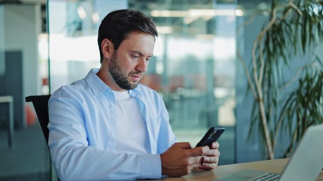 Stressed bearded businessman wearing blue shirt looking worried while reading bad news on smartphone in modern office, experiencing severe financial trouble and corporate pressure.