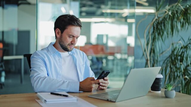 Stressed businessman, software developer wearing light blue shirt looking worried while checking smartphone in modern office environment, feeling concerned about bad news