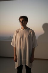 Obraz premium A young man stands in a serene indoor space, wearing an oversized white T-shirt and black pants, gazing out at a warm gradient sky through round glasses.