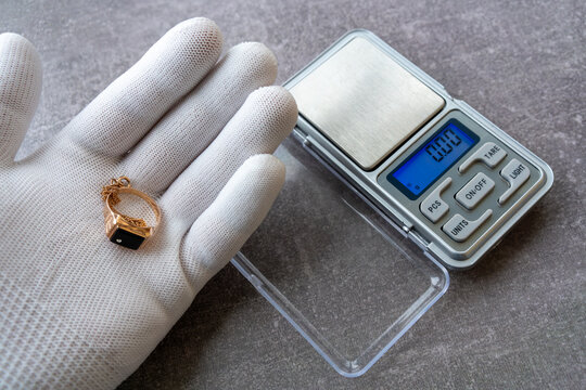 Hand in white glove holding gold ring with onyx stone near digital pocket scale. Jewelry appraisal and weighing of precious metal items.