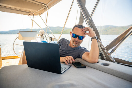 Relaxed man using laptop and smartphone while lounging on a sailing yacht. Digital nomad lifestyle and work life balance at sea.