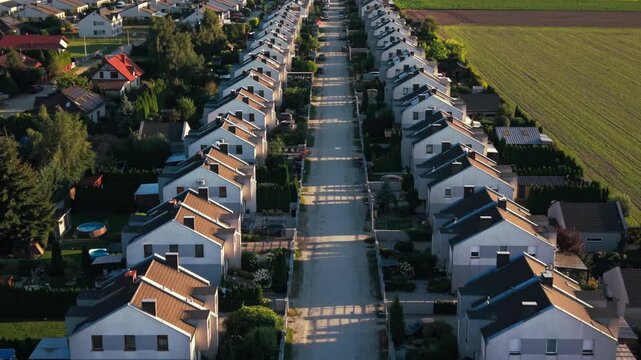 Aerial drone suburban residential neighborhood houses. This high-angle perspective showcases identical modern suburban homes arranged in neat rows along a central street, surrounded by green fields