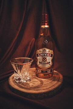 A bottle and an empty whiskey glass, Chivas Regal, stand on a wooden surface, with a dark background, there are no people