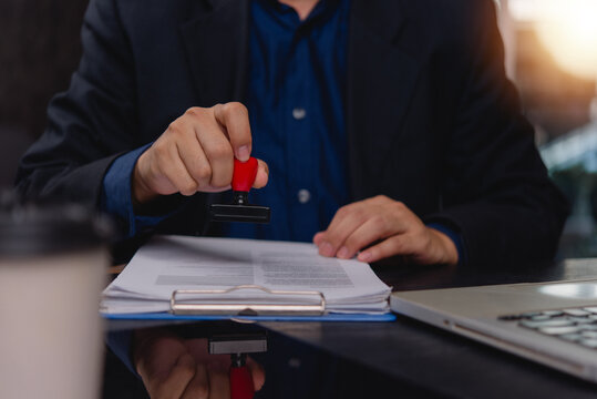 Business person stamping approval on document at office desk with laptop and clipboard for compliance verification and administrative workflow
