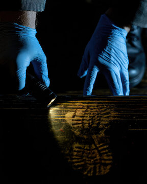 Forensic investigator in blue gloves examining faint bloody shoeprint on dark wooden stair with angled flashlight, revealing tread detail under high contrast crime scene lighting