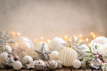 Naklejka premium A Christmas background featuring elegant white and silver baubles, glittering stars, jingle bells, and snowflakes on rustic wood, set against a warm backdrop with twinkling lights and copy space.